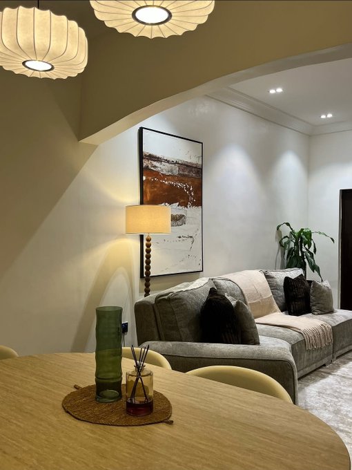 An interior photograph of a modern, stylish living room with a gray sofa, decorative pendant lights, an abstract painting, a table lamp, and a wooden dining table in the foreground.