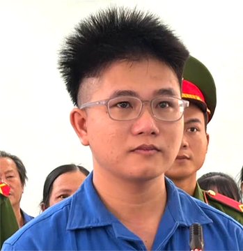 A young man with spiky hair and glasses, wearing a blue shirt, is shown in what appears to be a court or official setting, with uniformed personnel visible in the background. The image relates to the post about Nguyen Duy Nhat, a doctor from Hue Central Hospital who allegedly stole over 5 billion VND worth of gold to cover losses from failed crypto trading.