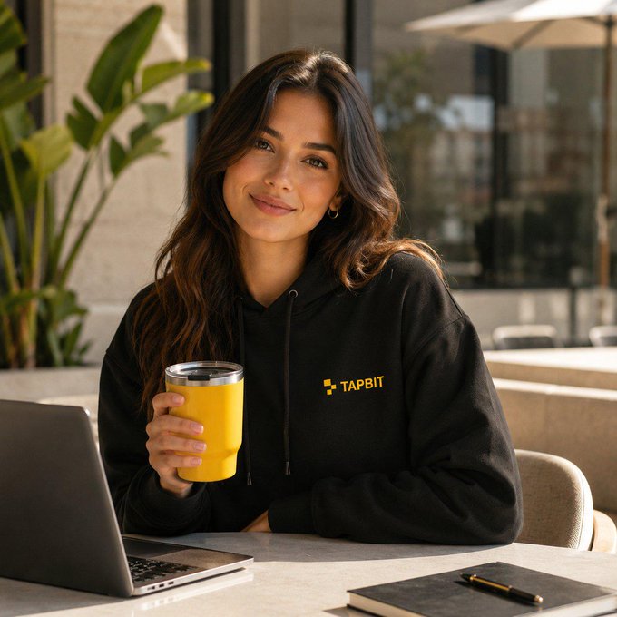 A smiling woman with long brown hair sits at an outdoor cafe table holding a yellow tumbler, wearing a black hoodie with 'TAPBIT' logo.