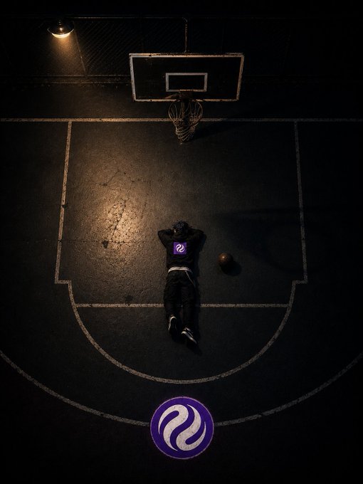 The image shows a person lying down on an indoor basketball court at night. The person appears to be resting or taking a break from playing basketball. The scene takes place within a dark room, which contrasts with the bright lights illuminating the basketball court.