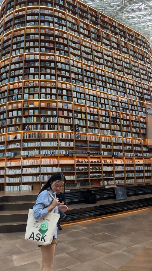 A young woman holding an 'ASK BOOK GROUP' tote bag stands inside a striking multi-story library with towering curved bookshelves filled with books.