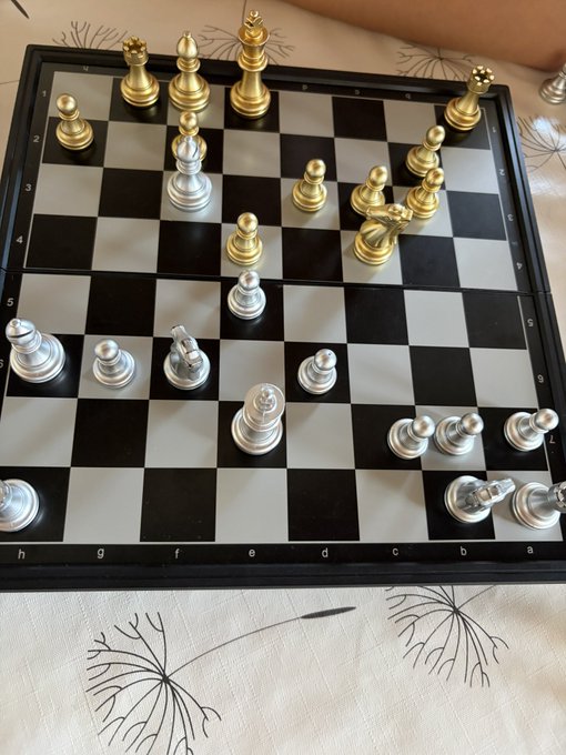A chess game in progress on a standard black and white board, with silver and gold pieces positioned mid-game on a floral-patterned tablecloth.