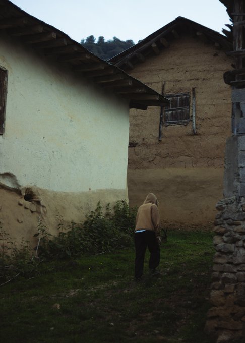 A person in a beige hoodie stands in the corner of two weathered, earthen-walled buildings in a rural setting.