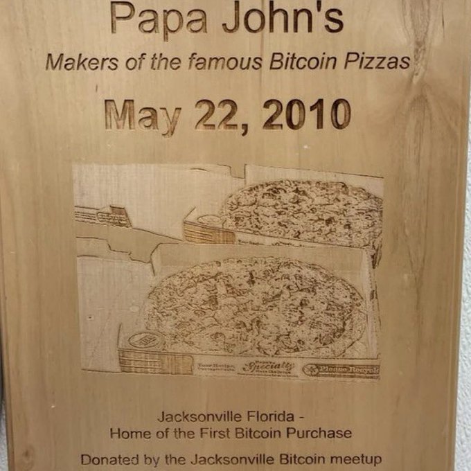 A wooden plaque commemorating 'Papa John's - Makers of the famous Bitcoin Pizzas - May 22, 2010' featuring an engraved illustration of two pizzas, noting Jacksonville Florida as the 'Home of the First Bitcoin Purchase.'
