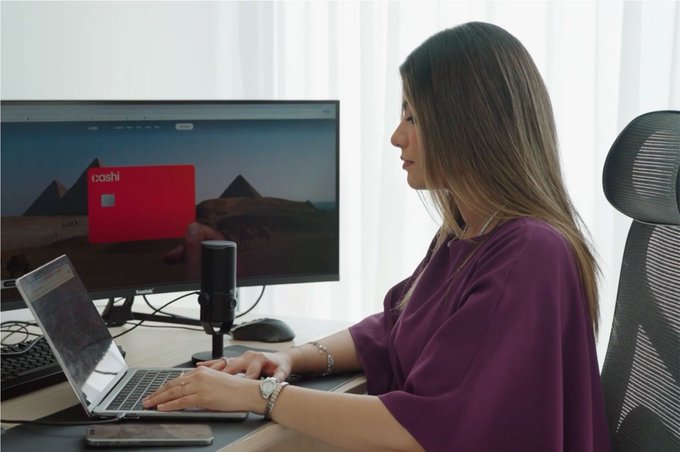 A Venezuelan professional based in the UAE sits at a sleek desk working on a laptop, with a red "cashl" branded card displayed on her monitor reflecting her fintech expertise. The setup, complete with a microphone, suggests content creation as she introduces her 10 years of experience in fintech, web3, and leadership roles at Binance and BNB Chain.