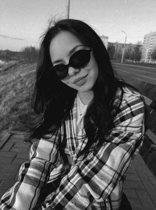 A black and white portrait of a young woman wearing sunglasses and a plaid shirt, posing outdoors with apartment buildings visible in the background.