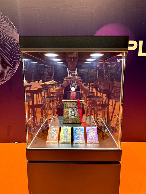 A glass display case containing a decorative object resembling a book and several colorful cards, with the reflection of a person taking a photo with a smartphone visible in the glass.