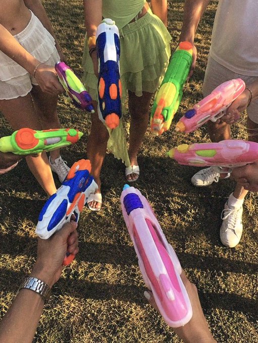 A top-down view of several hands holding colorful water guns pointed toward the center over a grassy surface, with people's legs visible around the edges.