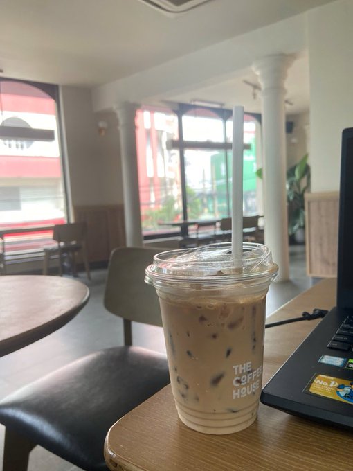 An iced coffee from 'THE COFFEE HOUSE' sits on a table next to a laptop, with large windows showing a red and white building outside.