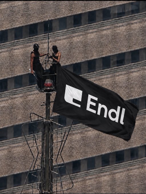 Two masked individuals in black sit atop a tower, deploying a large black flag with the Endrunde (Enda) party logo, a white stylized 'E'.