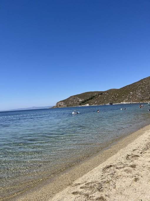 A scenic beach with clear turquoise water, sandy shore, and a mountainous coastline under a bright blue sky, with a few swimmers in the distance.