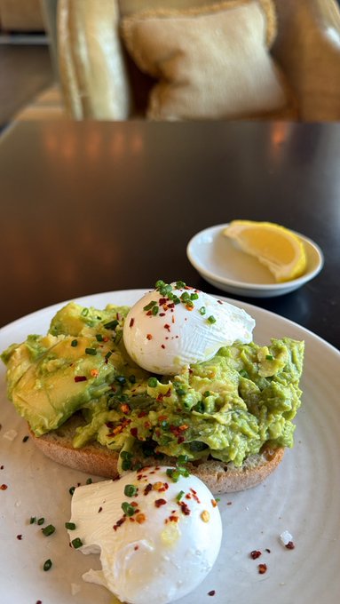 A beautifully plated avocado toast topped with poached eggs, chives, and red pepper flakes, served with a lemon wedge on the side—perfect for a satisfying "GM. Breakfast time" start to the day.