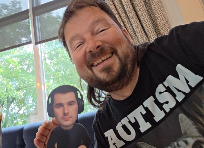 A smiling bearded man wearing a black "AUTISM" t-shirt holds up a small cutout photo of a man with headphones, posing in front of a bright window while sharing a cheerful "GM World" morning greeting and celebrating cooler 28°C weather in England compared to a previous 38°C scorcher.