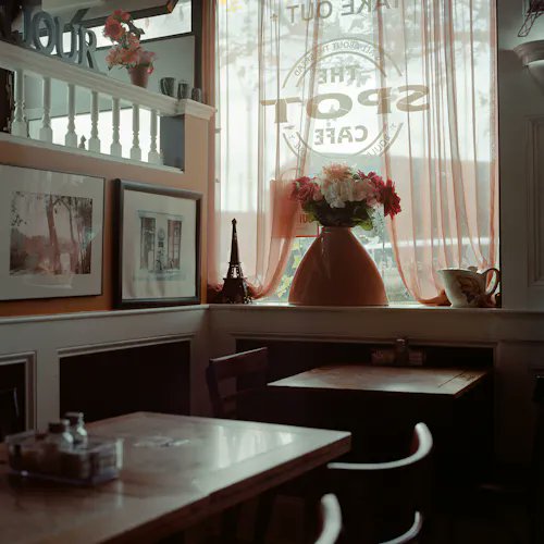 A cozy, dimly lit café interior featuring wooden tables, framed pictures on the walls, and a window with sheer curtains, a coffee shop sign, and a vase of pink flowers on the sill.