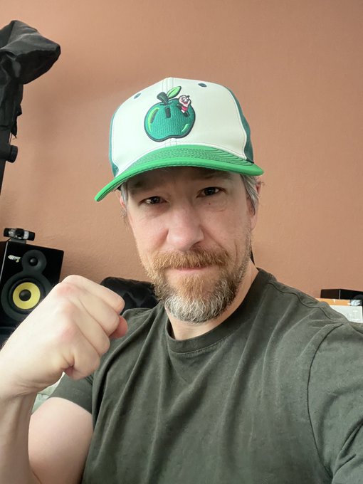 A bearded man wearing a green and white apple-themed baseball cap poses with a raised fist, with a yellow studio monitor visible in the background.