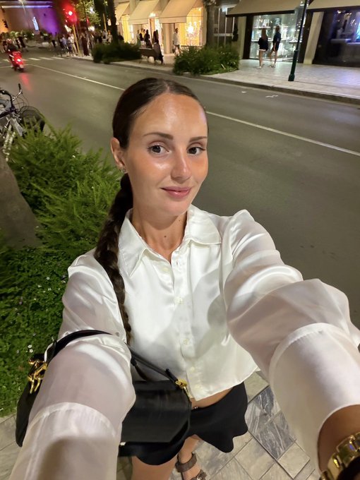A woman with a long brunette braid wearing a white button-up shirt takes a selfie on a city street at night, carrying a black handbag.