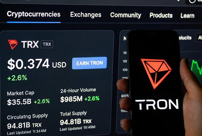 The image shows a person holding up their phone displaying a cryptocurrency trading app, which has a red logo for "TRON" on its screen. The person's hand is positioned over the phone, with the device held at an angle that allows them to see the screen clearly. The background of the image features a computer monitor or TV screen showing various data and information related to cryptocurrency exchanges.