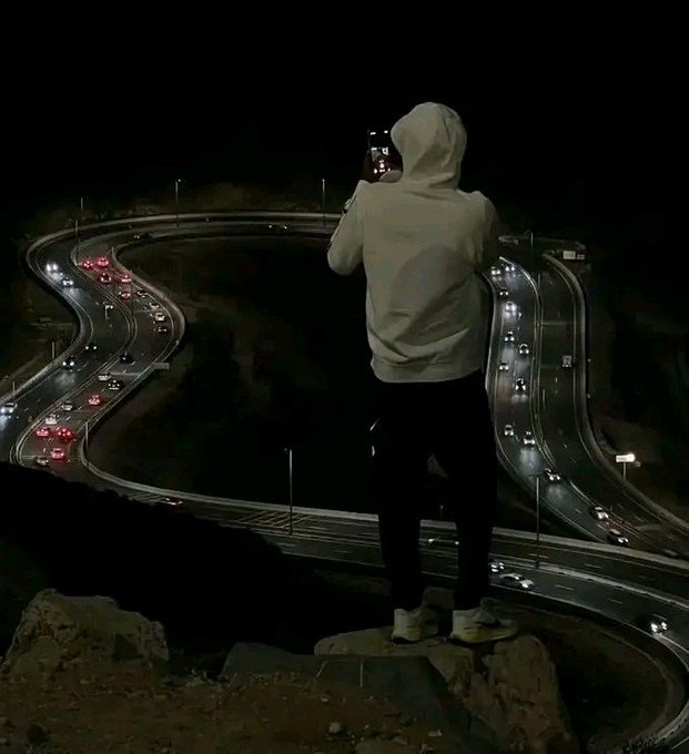 A person in a white hoodie stands on a rocky ledge overlooking a winding highway at night, capturing the winding trails of car lights below with their phone. The image evokes the "good night" and "see you at sunrise" sentiment from the post, with the illuminated roadway symbolizing the watchful eye being kept on $ANSEM overnight.