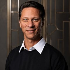 A professional headshot of a smiling man with dark hair, wearing a black sweater over a white collared shirt against a dark patterned background.