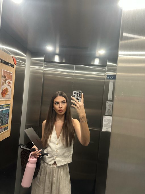 A woman with long brown hair takes a mirror selfie in an elevator while holding a pink water bottle, a laptop, and a phone with a decorative case.