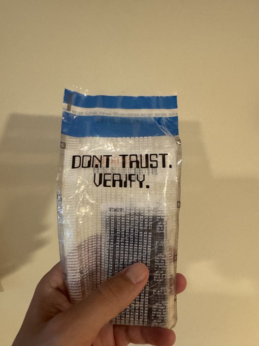 A hand holds a sealed clear plastic packet containing small components, printed with the text "DON'T TRUST. VERIFY."