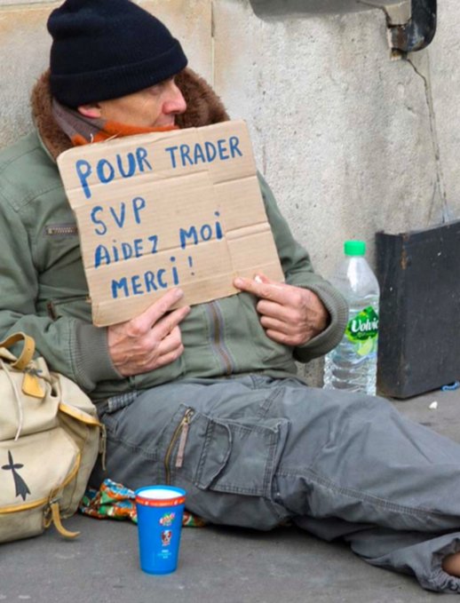 A homeless man sitting on the ground wearing a black beanie and green jacket, holding a cardboard sign in French that reads 'POUR TRADER SVP AIDEZ MOI MERCI !' (To trade please help me thank you!), with a bottle of Volvic water and a blue cup beside him.