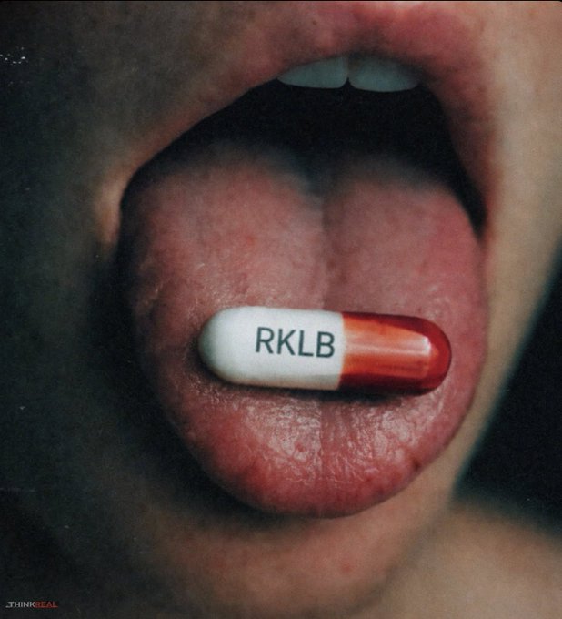 A close-up photograph of a person's mouth with their tongue extended, holding a red and white capsule pill with "RKLB" printed on it.
