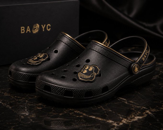 Black Crocs shoes featuring a gold skull charm and 'BAYC' branding, displayed next to a matching black box with gold logo.