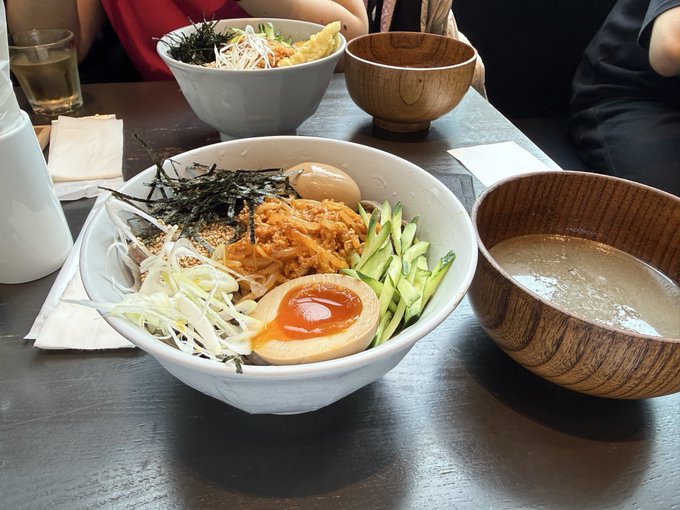 A table displays a Japanese tsukemen meal with a large bowl of noodles topped with a soft-boiled egg, kimchi, cucumber, and nori, accompanied by a wooden bowl of dipping broth and side dishes.