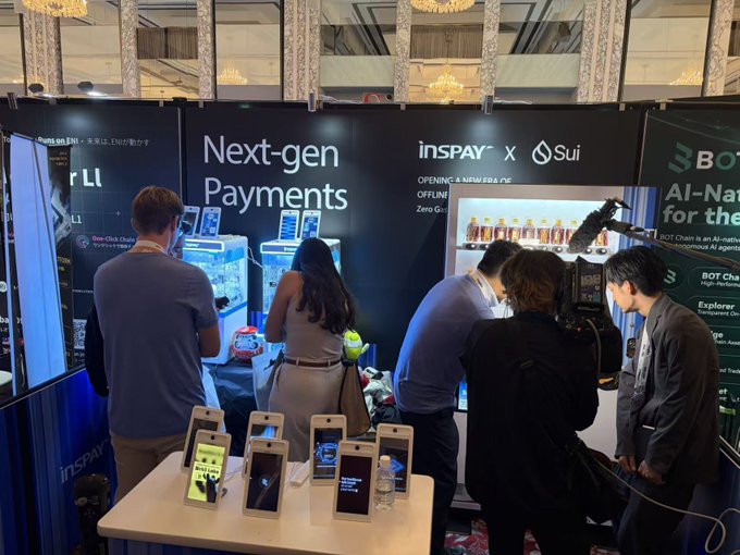 A convention booth scene with a large banner reading "Next-gen Payments" featuring "insPAY x Sui" branding. Several attendees interact with the display, which showcases payment devices and a vending machine, while a camera operator films the area.