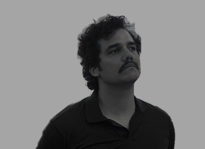 A black-and-white portrait of a man with dark curly hair and a mustache, looking upward against a plain gray background.