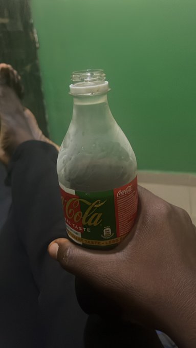 A hand holding a glass Coca-Cola bottle with a red label against a green wall background.