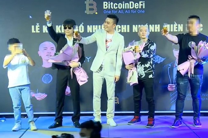 A photograph of an event for 'BitcoinDeFi - One for All, All for One' with Vietnamese text 'LỄ KÝ KẾT BỔ NHIỆM CHỈ NHÁNH MIỀN BẮC' on the backdrop, showing several men on stage holding champagne glasses and bouquets.