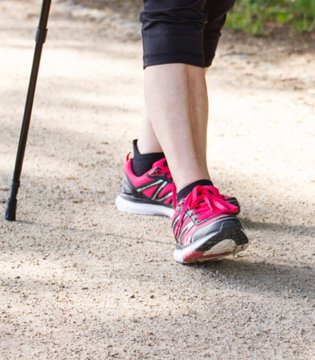 A person's legs in black shorts and pink running shoes walking on a dirt path with a walking pole visible on the left side.