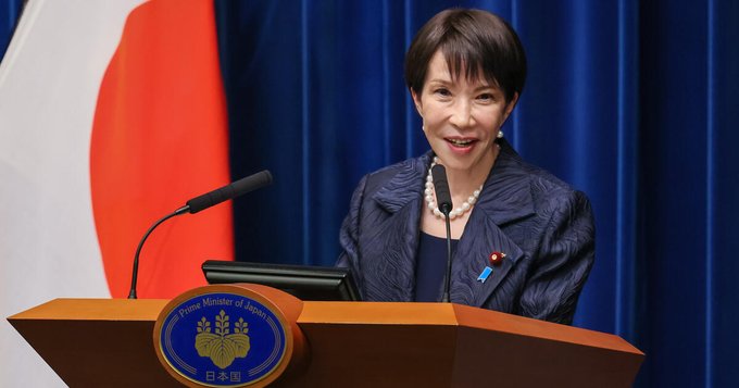 A woman stands at a podium bearing the emblem of the Prime Minister of Japan, positioned between the Japanese flag and a blue curtain. This image serves as a visual representation of the Bank of Japan (BOJ) and Japanese economic policy, providing context for the post's discussion on potential interest rate hikes and their impact on the Yen and cryptocurrency markets.