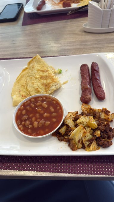 A breakfast plate on a wooden table containing baked beans in tomato sauce, an omelet, two sausages, and a mix of roasted potatoes and meat, with a phone and condiment holder in the background.