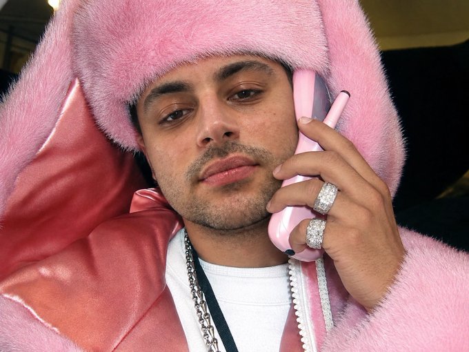 A young man in a pink fuzzy hoodie holding a pink telephone receiver to his ear, wearing a large diamond ring and a silver chain necklace.