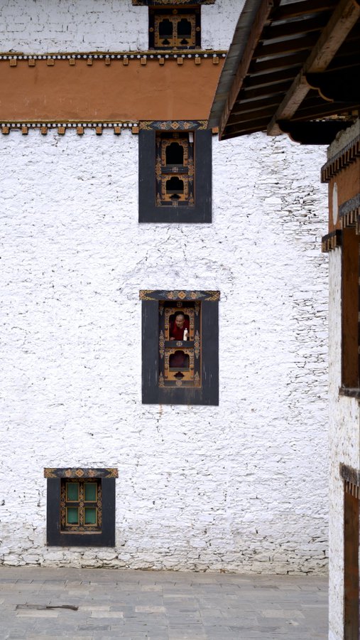 A photograph of a traditional whitewashed building, likely Bhutanese architecture, featuring intricately decorated dark wooden windows, orange trim, and a person wearing a red robe visible in the upper middle window.