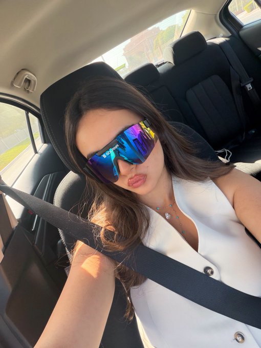 A young woman wearing mirrored Oakley sunglasses and a white top takes a selfie while sitting in the passenger seat of a car.