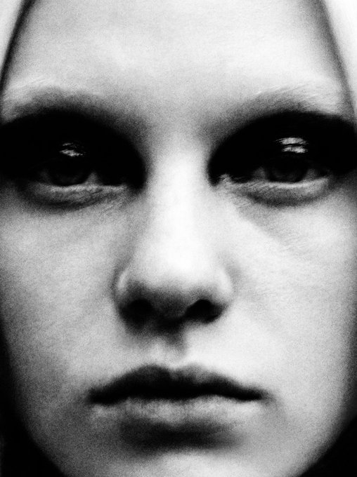 A high-contrast black and white close-up portrait focuses on a person's face with heavily shadowed eyes, creating a dramatic and intense expression.