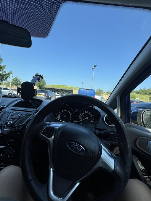 View from the driver's seat of a Ford (visible on the steering wheel) at a motorway service station, with the steering wheel and dashboard in the foreground and other cars and a clear blue sky visible through the windshield. The image captures the moment of a pit stop on the way to the Lake District, as referenced in the post where the traveler made stock purchases ($SIVE, $SMH) at the services rather than buying food.