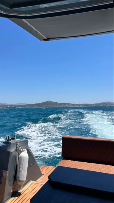 A view from the back of a boat traveling across bright blue ocean waters, with a coastline and hills visible in the distance under a clear sky.