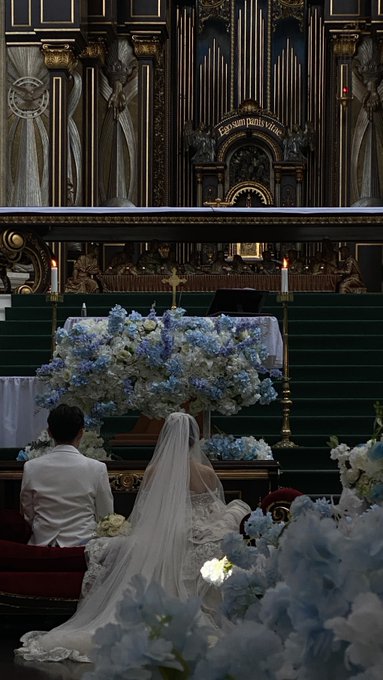 A wedding scene inside a grand church with ornate pipe organs, showing a bride in a long white veil and groom in a white suit kneeling before an altar adorned with blue and white floral arrangements.