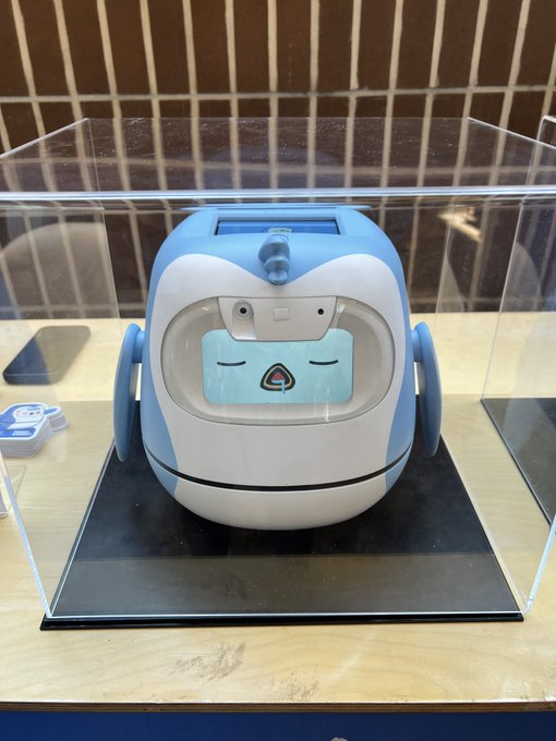 A blue and white cartoon-style robot with a sleepy face displayed on a screen sits inside a clear acrylic display case on a wooden table.