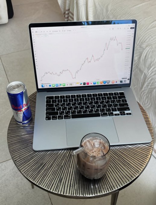 A workspace on a round wooden side table featuring a MacBook displaying a TradingView chart of an upward-trending stock, accompanied by a can of Red Bull and a glass of iced coffee.