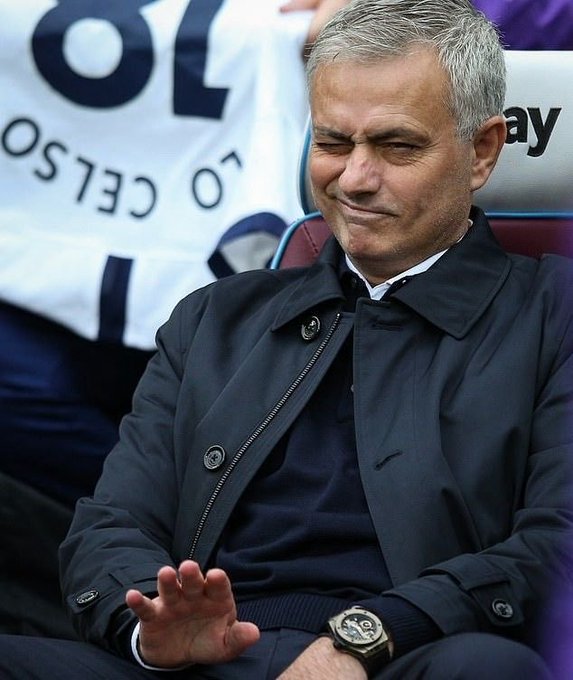 A man with gray hair in a dark coat sitting on a bench, making a displeased facial expression while gesturing with his hand.