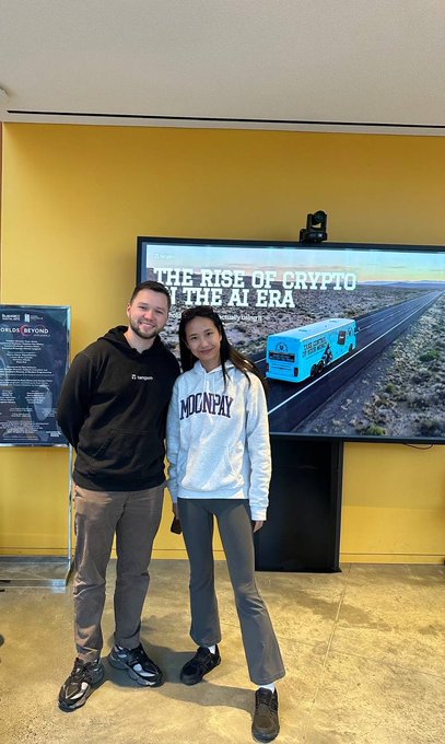 A young man and woman pose together in front of a screen displaying "THE RISE OF CRYPTO IN THE AI ERA" with a Bitcoin-themed truck on a highway, illustrating the message that "Crypto can be simple!"