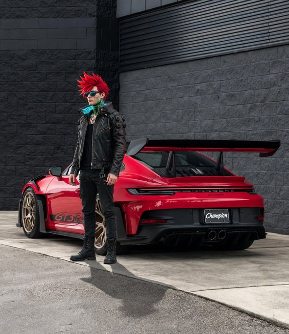 The image features a man with red hair standing next to a red sports car parked against a brick wall. He appears to be posing for a photo or video, possibly promoting the car. The focus of the content creation should be on showcasing the car's design, performance, and features rather than just the number of likes or followers. This approach can help create engaging content that appeals to a broader audience interested in the car itself, rather than solely focusing on social media metrics like likes and follows.