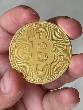 A close-up shot shows a hand holding a gold-colored physical Bitcoin commemorative coin with the 'B' logo and circuit-like engravings.