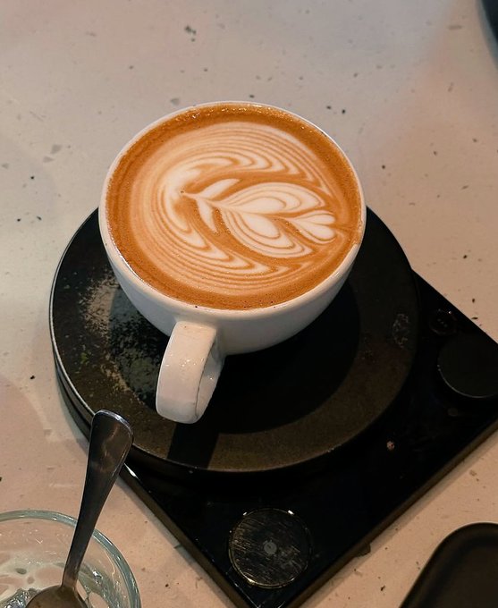 A flat white coffee with latte art sits on a black saucer atop a digital scale, with a spoon visible nearby. The image pairs with the "Gm gm Happy Sunday" post to convey a relaxed morning coffee moment while checking out the ARCTERMINAL AI platform.